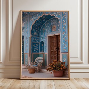 May include: Framed print of an ornate teal archway and doorway with floral patterns. A wicker chair and basket sit on a patio, with a terracotta pot of orange flowers. The print is in a wooden frame.