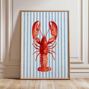 Puede incluir: Un estampado enmarcado con una ilustración de langosta roja sobre un fondo de rayas azules y blancas. La langosta está detallada con pinzas, patas y segmentos corporales. La obra de arte está presentada en un marco de madera.