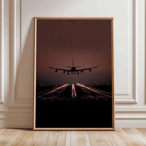 Könnte beinhalten: Gerahmter Druck eines Flugzeugs, das sich im Abenddämmerung einer Landebahn nähert. Die Silhouette des Flugzeugs steht vor einem dunkelroten Himmel, wobei die Landebahnlichter einen hellen Pfad bilden. Der Druck ist in einem Holzrahmen.