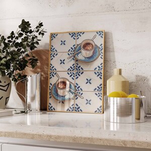 May include: A framed print of two cups of coffee on blue and white patterned tiles. The print is in a gold frame and is sitting on a white countertop.