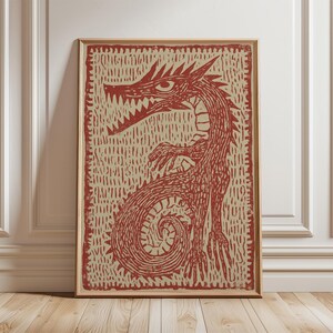 May include: A framed print featuring a red dragon illustration. The dragon has sharp teeth, a long neck, and a curled tail. The artwork is set against a textured beige background and framed in a wooden frame. The print is displayed on a wooden floor.