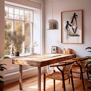 Op de afbeelding: Een lichte, luchtige kamer met een houten bureau en twee stoelen. Een ingelijst kunstwerk van een pumps hangt aan de muur. Een groot raam biedt uitzicht op bomen. Een zilveren hanglamp hangt boven het bureau.