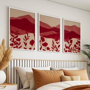 May include: Three framed art prints featuring abstract mountain landscapes in shades of red and beige. The artwork includes red floral elements at the bottom. The prints are displayed above a bed with orange and white pillows, creating a warm and inviting atmosphere.