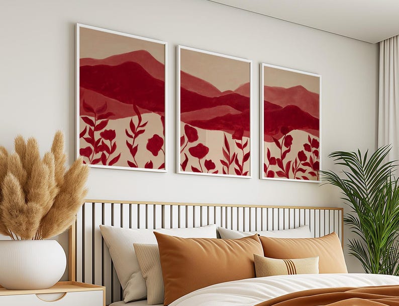 May include: Three framed art prints featuring abstract mountain landscapes in shades of red and beige. The artwork includes red floral elements at the bottom. The prints are displayed above a bed with orange and white pillows, creating a warm and inviting atmosphere.