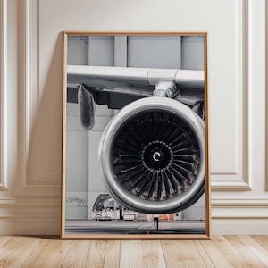 Könnte beinhalten: Gerahmter Druck mit einer Nahaufnahme eines Flugzeugtriebwerks. Das Triebwerk ist silberfarben mit schwarzen Lüfterblättern, vor einem grauen Hangarhintergrund. Eine Person ist in der Ferne zu sehen. Der Druck ist in einem Holzrahmen.