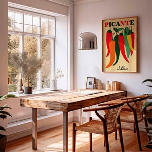 May include: A dining room scene featuring a wooden table with metal legs and two woven chairs. A framed poster with the word "PICANTE" and colorful chili peppers hangs on the wall. A large window offers a view of trees.