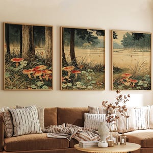 May include: Three framed art prints showing a forest scene with red and white mushrooms. The artwork features tall trees, a body of water, and lush greenery. The prints are displayed above a brown sofa with striped cushions.