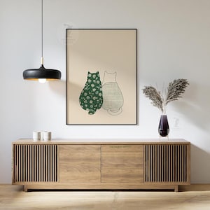 Puede incluir: Una impresi&oacute;n art&iacute;stica enmarcada con dos gatos estilizados. Uno es verde con un estampado floral blanco, el otro es un contorno simple. La impresi&oacute;n cuelga sobre un mueble de madera con detalles de listones verticales. Una l&aacute;mpara colgante negra cuelga del techo.