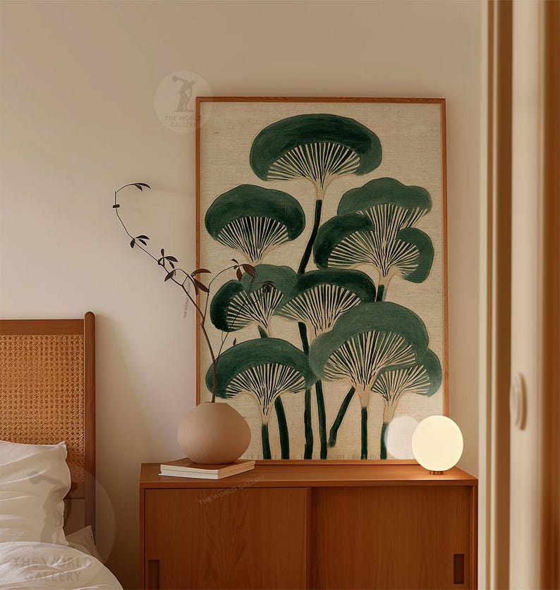 May include: A framed botanical print featuring stylized green mushroom illustrations on a beige background. The artwork is displayed above a wooden cabinet with a round beige vase, a book, and a small, round lamp.