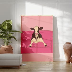 May include: A framed art print depicting a cow relaxing on a pink sofa. The cow is black and white with a pink nose. The background is a solid pink. The print is displayed in a room with a plant, ottoman, and decorative vases.