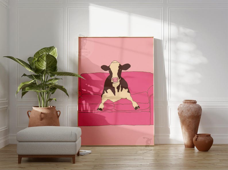 May include: A framed art print depicting a cow relaxing on a pink sofa. The cow is black and white with a pink nose. The background is a solid pink. The print is displayed in a room with a plant, ottoman, and decorative vases.