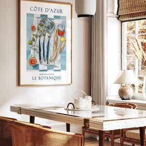 May include: A framed art print featuring a still life of fish, olives, bread, and citrus fruits on a blue and white checkered cloth. The print has the text "C&Ocirc;TE D'AZUR" and "LE BOTANIQUE". The print is displayed in a room with a table and chairs.