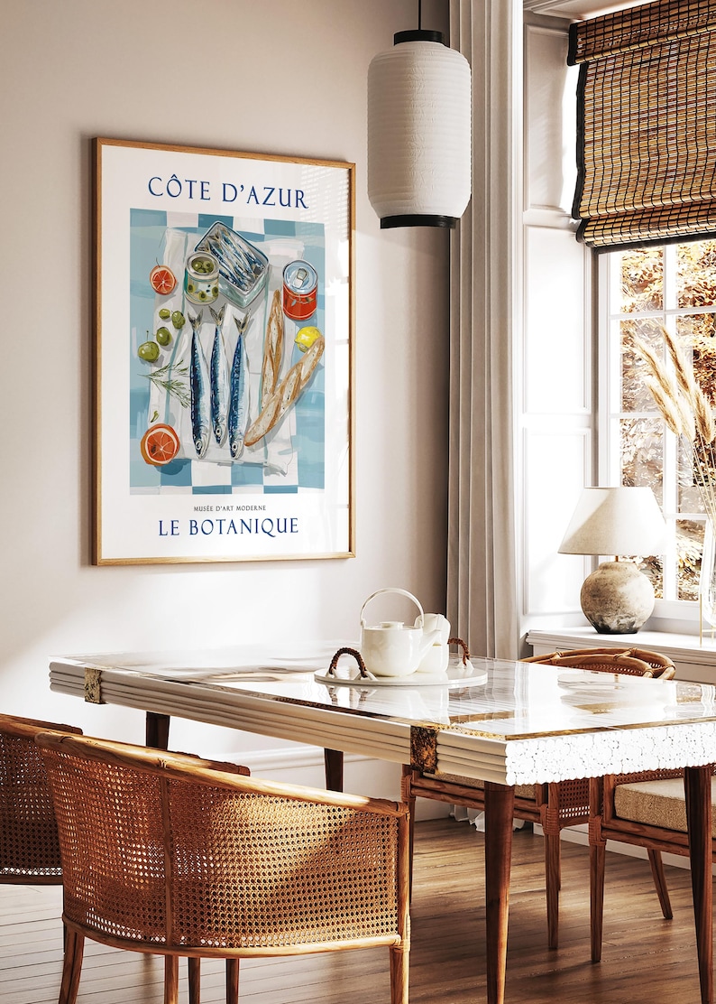 May include: A framed art print featuring a still life of fish, olives, bread, and citrus fruits on a blue and white checkered cloth. The print has the text "C&Ocirc;TE D'AZUR" and "LE BOTANIQUE". The print is displayed in a room with a table and chairs.
