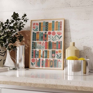 May include: A framed print of a colorful illustration featuring a wall of books with flowers and leaves growing from the spines. The print has a gold frame and is displayed on a white countertop.