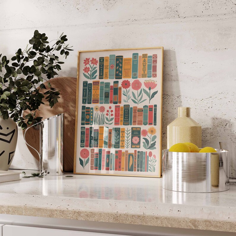 May include: A framed print of a colorful illustration featuring a wall of books with flowers and leaves growing from the spines. The print has a gold frame and is displayed on a white countertop.