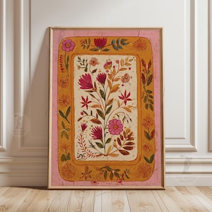 May include: A framed floral art print with a central display of pink and red flowers and green leaves. The artwork is set against a cream background, framed by a gold border and a pink outer frame. The print is displayed on a wooden floor.