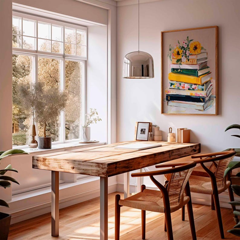 May include: A bright, airy room with a wooden desk and two chairs. A framed painting of a stack of books with sunflowers hangs on the wall. A large window offers a view of trees. A silver pendant light hangs above the desk.