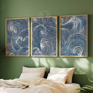 May include: Three framed wall art pieces with a blue and white wave pattern. The artwork is displayed on a green wall above a bed with white bedding. The frames are a light gold color.