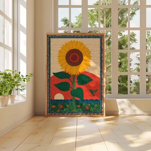Op de afbeelding: Een ingelijst kunstwerk met een grote gele zonnebloem met een bruin centrum, groene bladeren en een rode zonsondergang achtergrond. Een kolibrie is onderaan afgebeeld. Het kunstwerk wordt tentoongesteld in een kamer met een raam en planten.