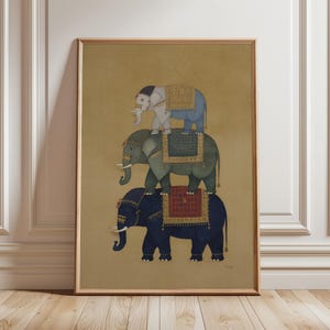 May include: Framed art print depicting three elephants, one atop the other. The elephants are painted in blue, green, and grey, with decorative saddle blankets in red, gold, and blue. The background is a muted gold colour.