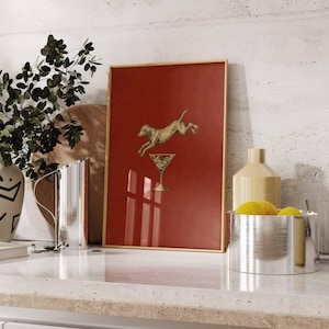 Pu&ograve; includere: Stampa artistica incorniciata con un cane dorato che salta sopra un bicchiere da martini su sfondo rosso. La stampa ha una cornice dorata ed &egrave; esposta su un piano di lavoro bianco con altri oggetti decorativi.