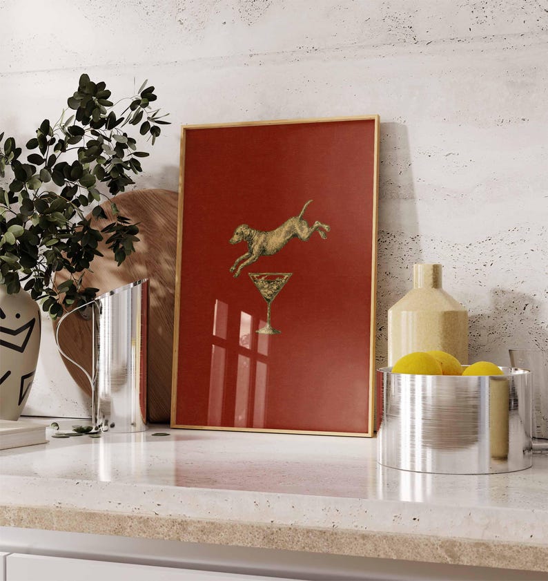 Pu&ograve; includere: Stampa artistica incorniciata con un cane dorato che salta sopra un bicchiere da martini su sfondo rosso. La stampa ha una cornice dorata ed &egrave; esposta su un piano di lavoro bianco con altri oggetti decorativi.