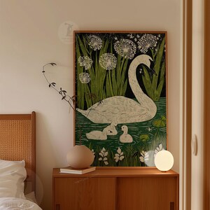 May include: A framed art print featuring a white swan with three cygnets in a pond, surrounded by green reeds and white flowers. The artwork has a black background and a wooden frame. The print is displayed on a wooden cabinet.