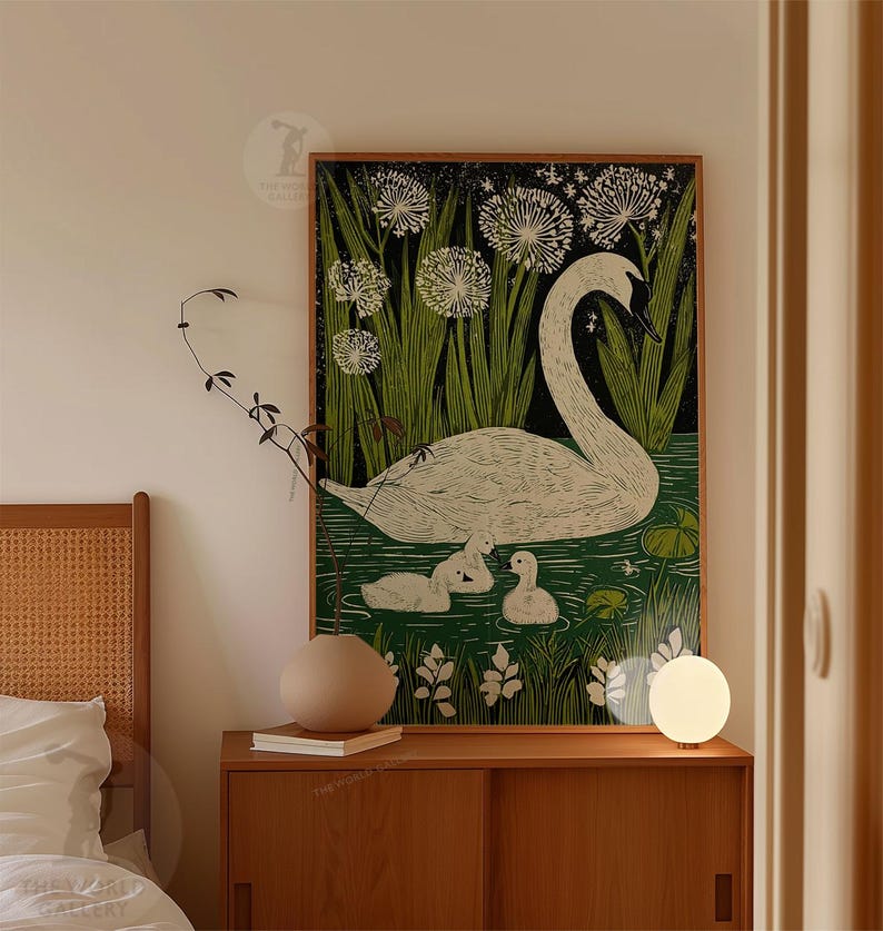 May include: A framed art print featuring a white swan with three cygnets in a pond, surrounded by green reeds and white flowers. The artwork has a black background and a wooden frame. The print is displayed on a wooden cabinet.