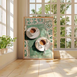 May include: A framed print featuring two cups of coffee on saucers, resting on a green and white patterned surface. The artwork is set against a backdrop of a sunlit room with a large window and wooden floor.