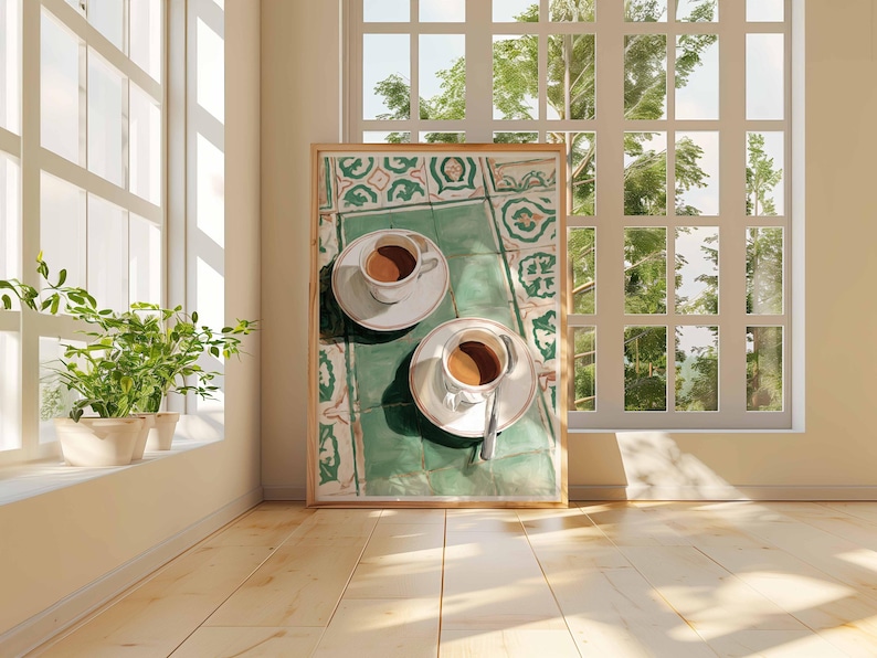 May include: A framed print featuring two cups of coffee on saucers, resting on a green and white patterned surface. The artwork is set against a backdrop of a sunlit room with a large window and wooden floor.