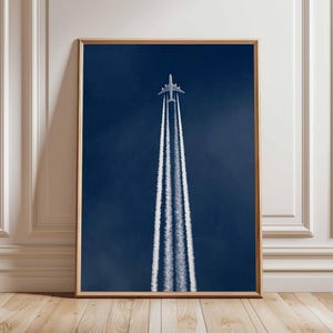 Op de afbeelding: Een ingelijste kunstdruk met een vliegtuig tegen een diepblauwe lucht. Het vliegtuig laat drie witte condensstrepen achter die zich naar de onderkant van het frame uitstrekken. De print zit in een houten lijst en staat tegen een witte muur.