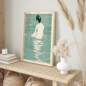 May include: A framed print of a woman with dark hair sitting in a pool of water. The water is a light blue-green color and the woman is facing away from the viewer. The print is in a wooden frame.