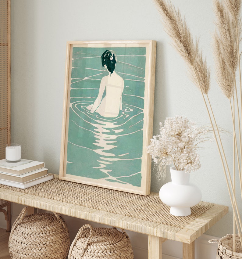 May include: A framed print of a woman with dark hair sitting in a pool of water. The water is a light blue-green color and the woman is facing away from the viewer. The print is in a wooden frame.