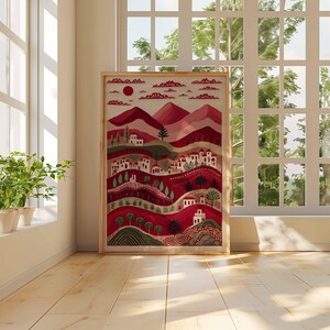 May include: A framed art print depicting a stylized landscape with red hills, white buildings, and trees. The artwork uses a warm colour palette with shades of red, green, and white. The print is displayed in a light-coloured wooden frame.