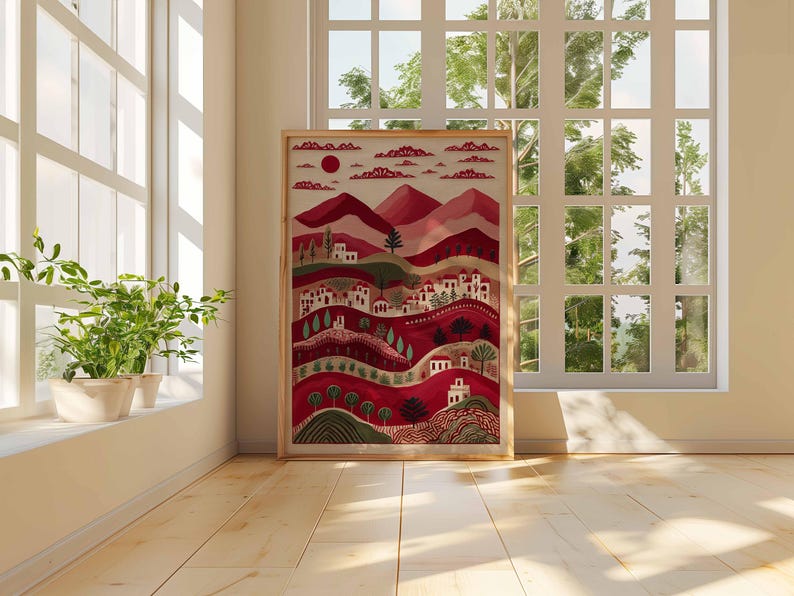 May include: A framed art print depicting a stylized landscape with red hills, white buildings, and trees. The artwork uses a warm colour palette with shades of red, green, and white. The print is displayed in a light-coloured wooden frame.