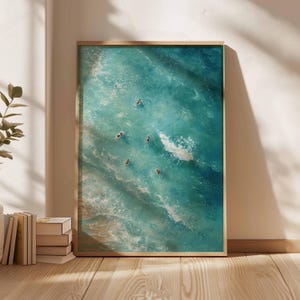 May include: A framed art print featuring an aerial view of several people swimming in turquoise ocean water. The artwork is displayed in a light-colored wooden frame, leaning against a white wall. The scene is illuminated by natural light.