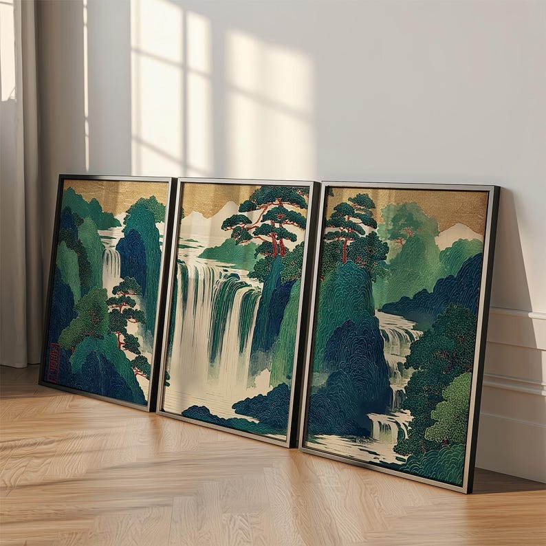 May include: Three framed art prints depicting a Japanese landscape with waterfalls, trees, and mountains. The artwork features a color palette of green, blue, and gold, with black frames. The prints are leaning against a wall on a wooden floor.
