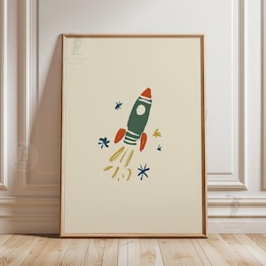 以下が含まれることがあります： ロケットの子供っぽいイラストが描かれたフレームプリント。ロケットは濃い緑色で、赤いアクセントと赤いノーズがあり、黄色の炎と青と黄色の星があります。アートワークはクリーム色の背景に設定されています。