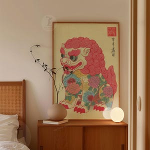 May include: A framed print of a colorful, stylized lion-dog with floral patterns. The artwork features a red mane, blue and pink floral accents, and traditional Asian script. The print is displayed on a wooden cabinet with a neutral-toned vase and a small lamp.