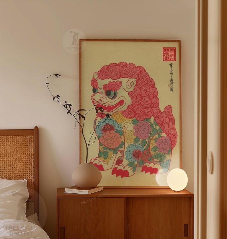 May include: A framed print of a colorful, stylized lion-dog with floral patterns. The artwork features a red mane, blue and pink floral accents, and traditional Asian script. The print is displayed on a wooden cabinet with a neutral-toned vase and a small lamp.
