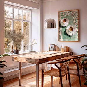 May include: A bright, airy interior with a wooden desk and two chairs. A framed artwork of two cups of coffee hangs on the wall. A large window offers a view of trees. A silver pendant light hangs above the desk.