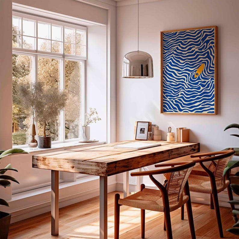 May include: A bright, airy room with a wooden desk and two chairs. A framed art print with a blue and white wave pattern and a yellow surfboard hangs on the wall. A large window offers a view of trees.
