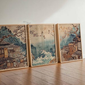 May include: Three framed prints of Japanese art depicting a traditional village with cherry blossoms and birds flying over a body of water. The prints are in a light wood frame and have a white background.