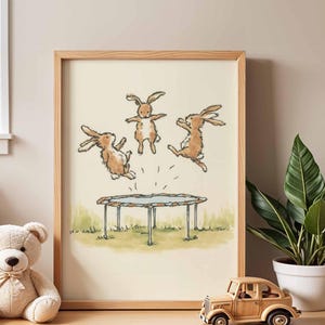 May include: A framed print featuring three brown rabbits jumping on a trampoline. The illustration has a light beige background with green grass at the bottom. The frame is made of light-colored wood.