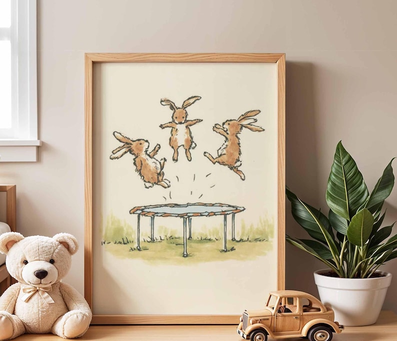 May include: A framed print featuring three brown rabbits jumping on a trampoline. The illustration has a light beige background with green grass at the bottom. The frame is made of light-colored wood.