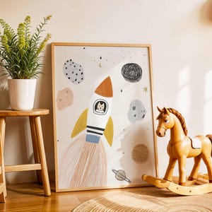May include: A framed art print featuring a cartoon rocket ship with a cat in the cockpit, surrounded by planets and stars. A wooden rocking horse and a potted plant are in the foreground. The art print is in a light wood frame.