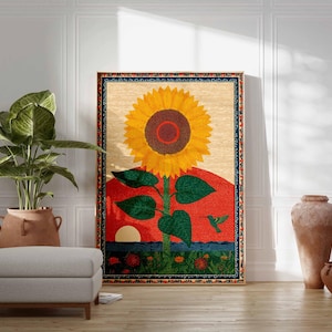 Op de afbeelding: Een groot ingelijst tapijt met een levendige zonnebloem tegen een rode en oranje zonsondergang. Het kunstwerk bevat een groene kolibrie en een blauwe rand. Een plant in pot, een poef en decoratieve vazen staan op de voorgrond.