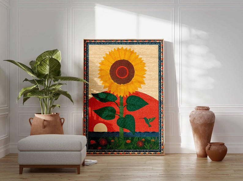 Op de afbeelding: Een groot ingelijst tapijt met een levendige zonnebloem tegen een rode en oranje zonsondergang. Het kunstwerk bevat een groene kolibrie en een blauwe rand. Een plant in pot, een poef en decoratieve vazen staan op de voorgrond.