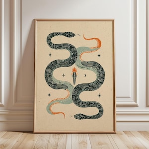 May include: A framed wall art print on a light beige background featuring two intertwined snakes in dark green, sage green, and orange. The design includes a central cactus illustration, small star symbols, and a crescent moon, all rendered in a textured, block-print style.