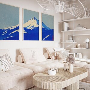 K&ouml;nnte beinhalten: Drei gerahmte Kunstdrucke, die eine Bergkette in Blau- und Wei&szlig;t&ouml;nen zeigen. Das Kunstwerk h&auml;ngt &uuml;ber einem hellfarbigen Ecksofa mit dekorativen Kissen. Ein Holztisch und ein wei&szlig;es Regal vervollst&auml;ndigen die Einrichtung des Raumes.